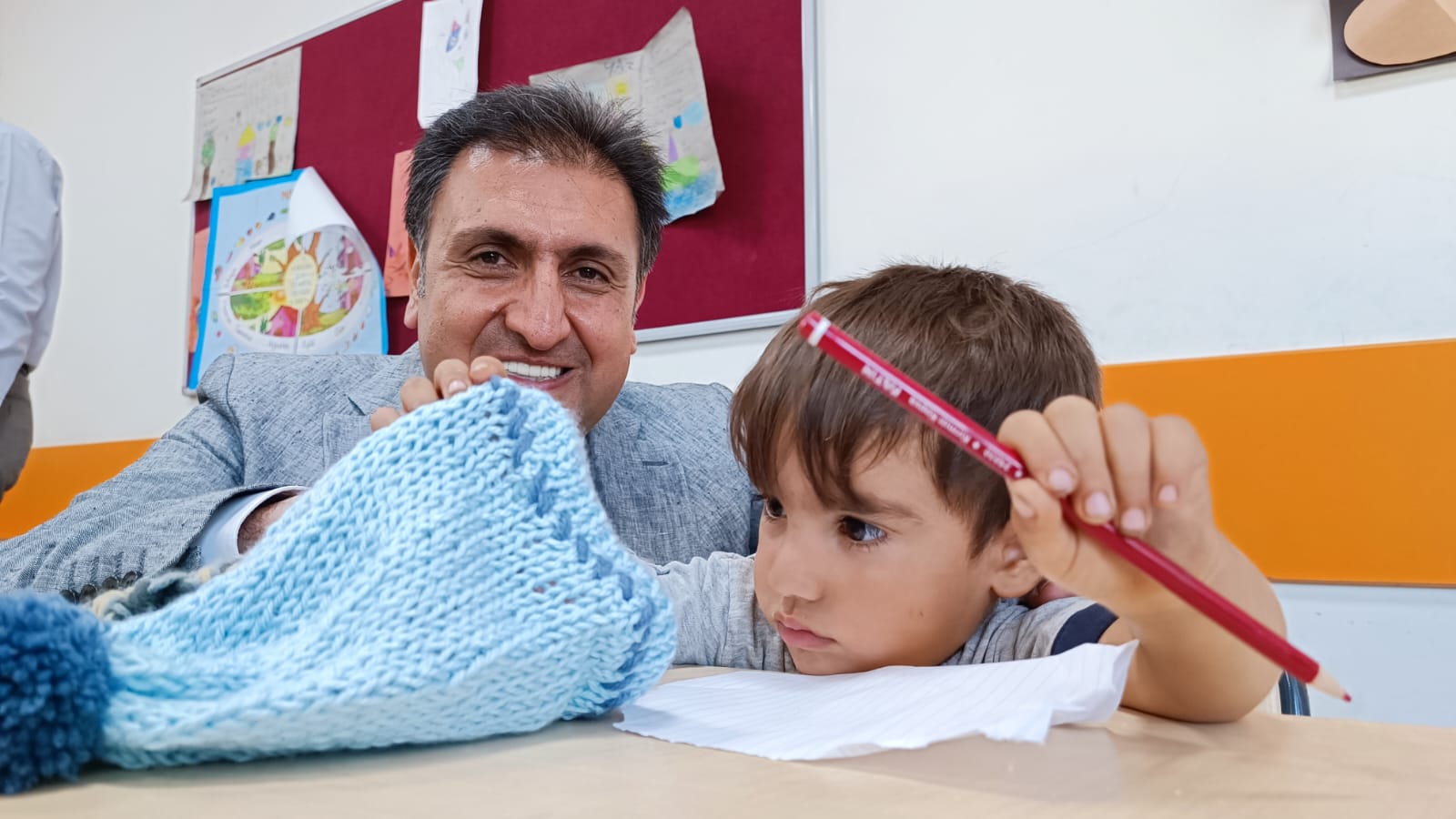 A Woolly Wishes volunteer smiling with a young boy at his school desk, a handmade blue knitted gift in front of them