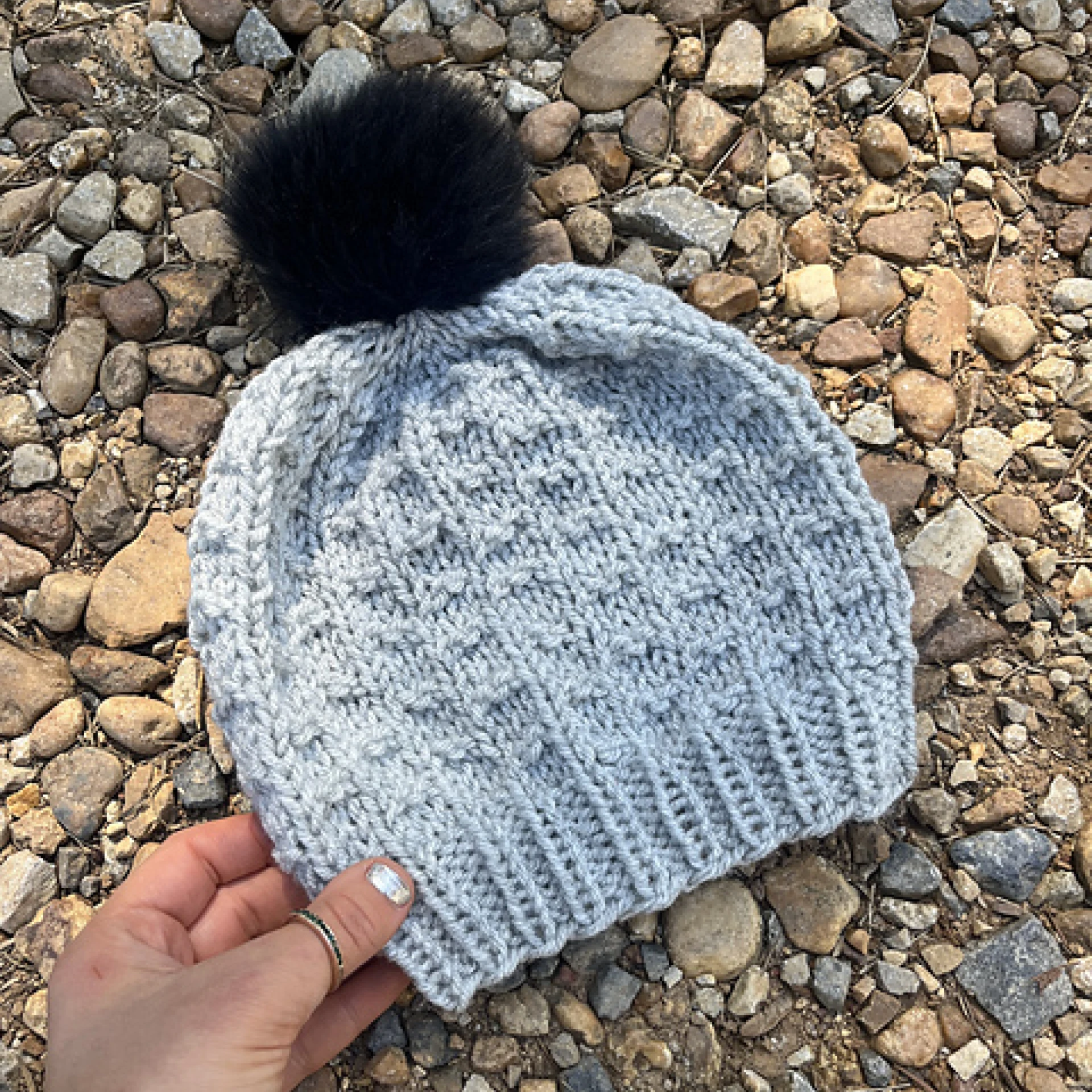 Light blue Wishes Beanie with a black pom-pom, laid on rocky ground