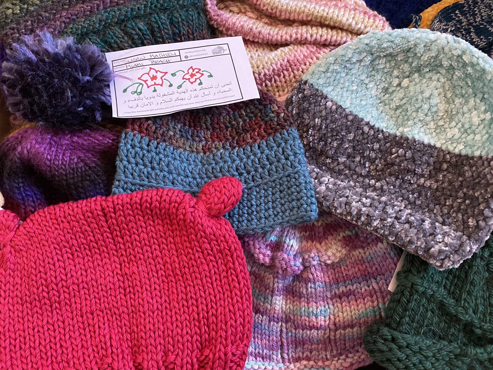 A pile of handmade knit hats in many colors, each with a maker's tag